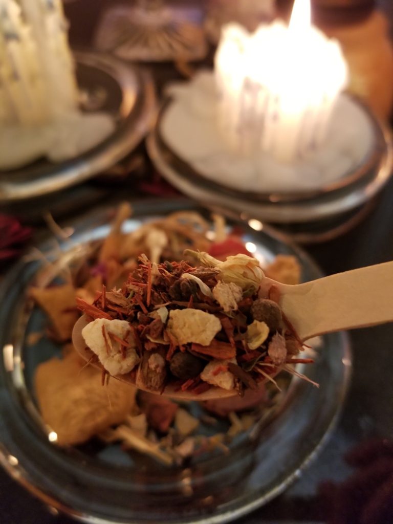 A wooden spoon with a mixture of dried herbs and a candle burning in the background