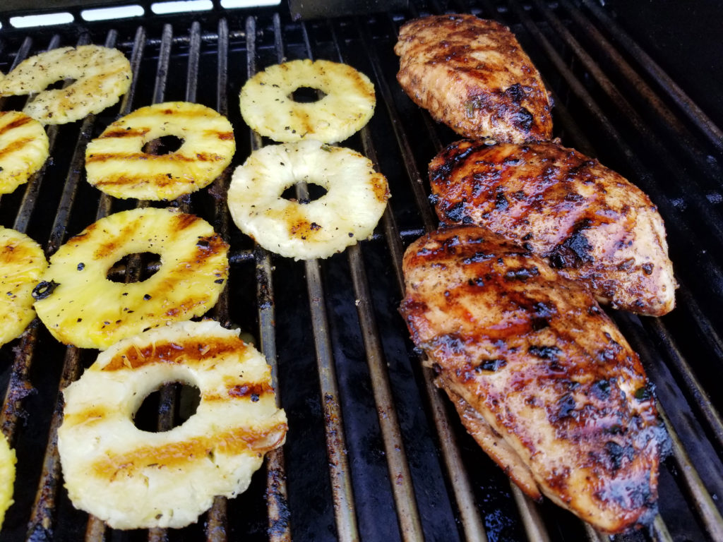 Pineapple rings on a grill with golden grill marks. Also on the grill is char broiled chicken breast with grill marks