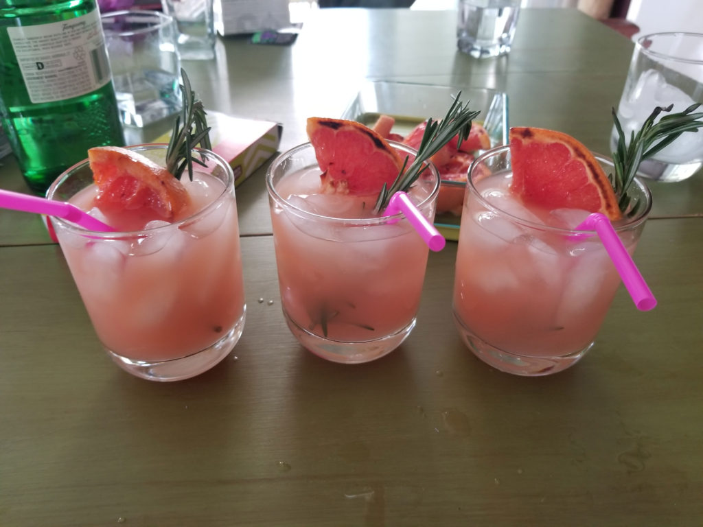 Three rocks glasses filled with ice and pink grapefruit juice. Garnished with a pink straw, a piece of grilled grapefruit and a sprig of rosemary. Sitting on a mossy green table