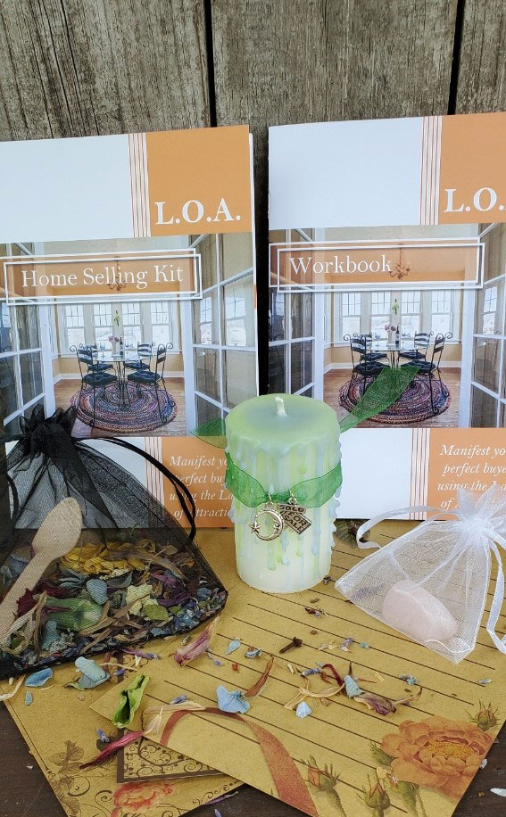 A photo of the LOA Home Selling whole kit. A workbook, Instruction book, polished crystal, dried herbs with a wooden spoon, a handmade white pillar candle with a light lime green wax drip Reiki charged candle wrapped in a green ribbon and charms, natural stationary,