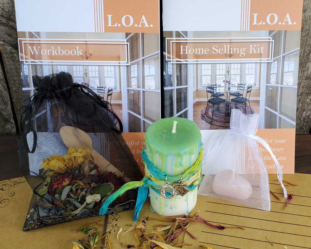 A photo of the LOA Home Selling whole kit. A workbook, Instruction book, polished crystal, dried herbs with a wooden spoon, a handmade Reiki charged white pillar candle with a light lime green and blue wax drip wrapped in a green and blue ombre ribbon and charms and natural stationary,