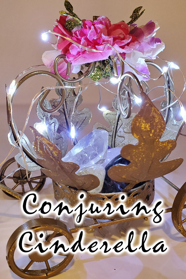 A small table top gold carriage with pink and purple flowers and lights holding a clear quartz crystal, rose quartz crystal black onyx crystal, amethyst and a citrine crystal with the words Conjuring Cinderella