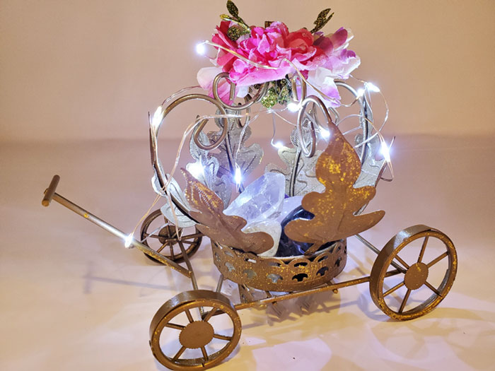 A small table top gold carriage with pink and purple flowers and lights holding a clear quartz crystal, rose quartz crystal black onyx crystal, amethyst and a citrine crystal