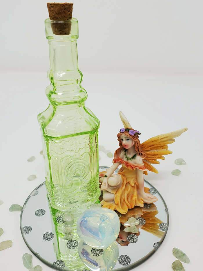 A lime green corked potion bottle, an opalite crystal and a sitting yellow fairy sitting on a round mirror with sparkly etched polka dots