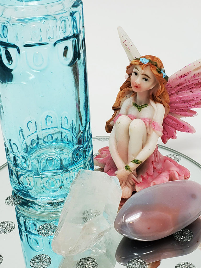 A clear quartz crystal and Botswana crystal sitting on a mirror with a blue bottle and pink sitting fairy with the words manifest with fairy rain and crystals