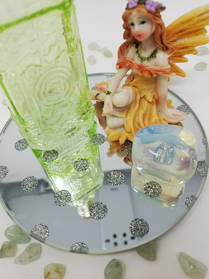 A lime green corked potion bottle, an opalite crystal and a sitting yellow fairy sitting on a round mirror with sparkly etched polka dots