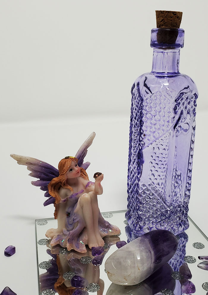 A purple sitting fairy with an amethyst crystal and a purple corked potion bottle sitting on top of a mirror