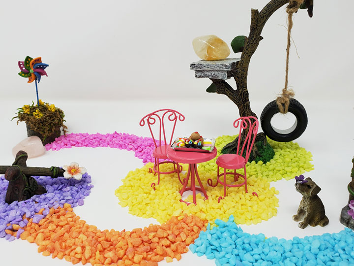 A colorful fairy garden scene representing a labyrinth or a medicine wheel. Colorful blue, yellow, orange, purple, pink & green stones create pathways. At the end of each path are fairy garden items such as a teeter totter, a fairy door, a mini dog, an owl, a tree swing, a pink bistro table and chairs with a desert tray on the table, a pinwheel and 3 crystals, rose quartz, clear quartz and citrine. A fairy garden for spiritual balance