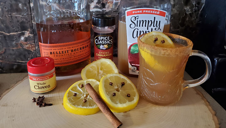 photo of ingredients to make a Hot Toddy drink. McCormick's Whole cloves, lemon wedges with the cloves stuck in, Spice Classics Cinnamon sticks, A bottle of Bulleit Bourbon, a bottle of Simply Apple Juice, and a cinnamon rimmed mug filled with a lemon & a finished Hot Toddy