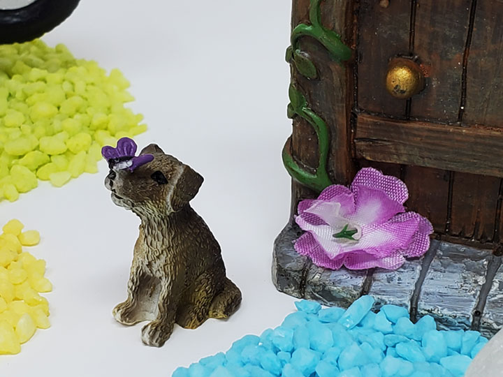 A mini dog with a purple butterfly on his nose sitting next to a fairy door with a purple flower on it