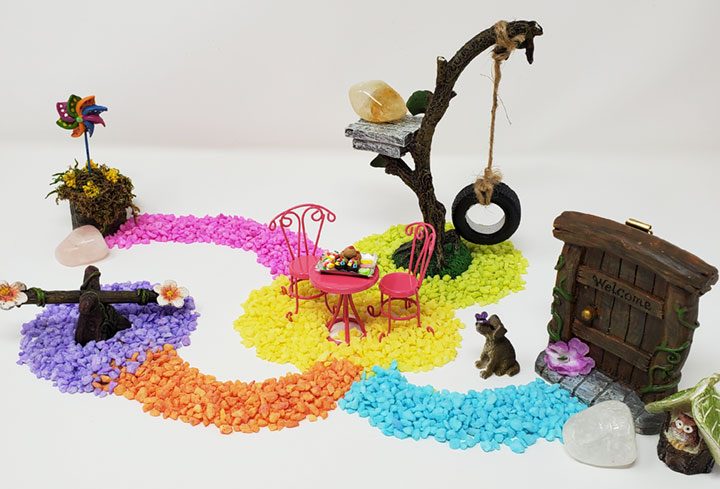 A colorful fairy garden scene representing a labyrinth or a medicine wheel. Colorful blue, yellow, orange, purple, pink & green stones create pathways. At the end of each path are fairy garden items such as a teeter totter, a fairy door, a mini dog, an owl, a tree swing, a pink bistro table and chairs with a desert tray on the table, a pinwheel and 3 crystals, rose quartz, clear quartz and citrine. A fairy garden for spiritual balance