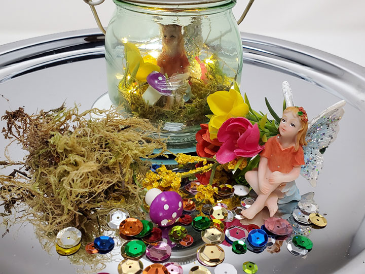 colorful shiny sequins with artificial yellow, red and pink flowers, dried moss, a purple mini mushroom and a finished fairy lights jar