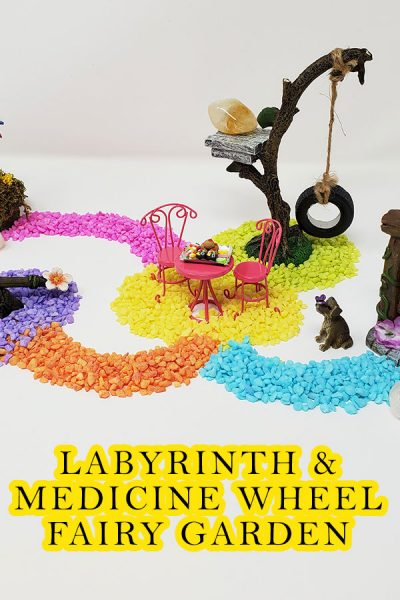 A colorful fairy garden scene representing a labyrinth or a medicine wheel. Colorful blue, yellow, orange, purple, pink & green stones create pathways. At the end of each path are fairy garden items such as a teeter totter, a fairy door, a mini dog, an owl, a tree swing, a pink bistro table and chairs with a desert tray on the table, a pinwheel and 3 crystals, rose quartz, clear quartz and citrine. A fairy garden for spiritual balance