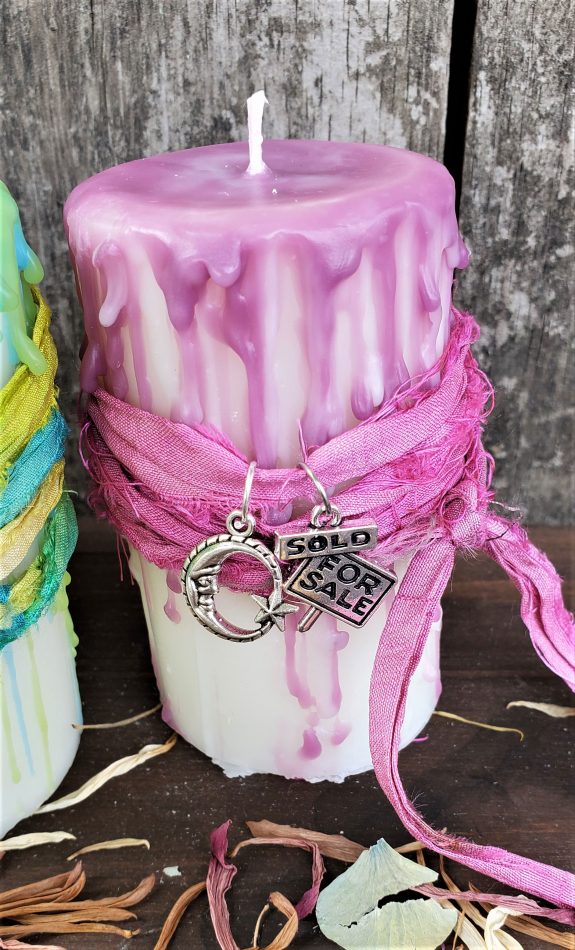 A handmade Reiki charged white pillar candle with fuschia color wax drip and ribbon with a For sale/sold charm and a moon charm